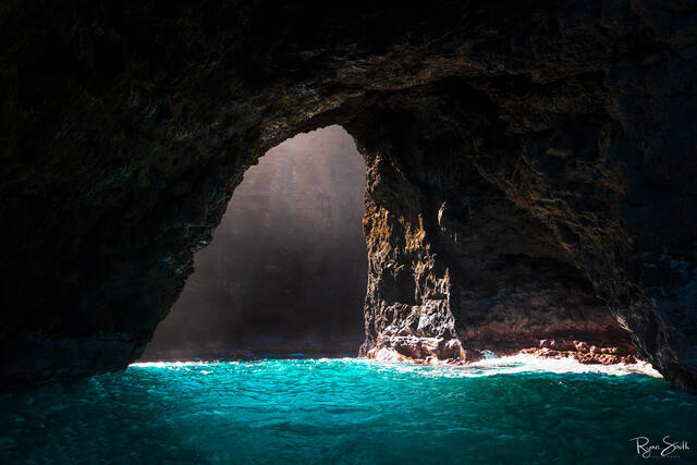Inside a sea cave, the sunlight shines in and lights the the turquoise water and the dark cave walls. 