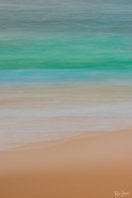 Vertical abstract photography of the beach close up with the tan sand, shallow water and deep turquoise and blue water out further.