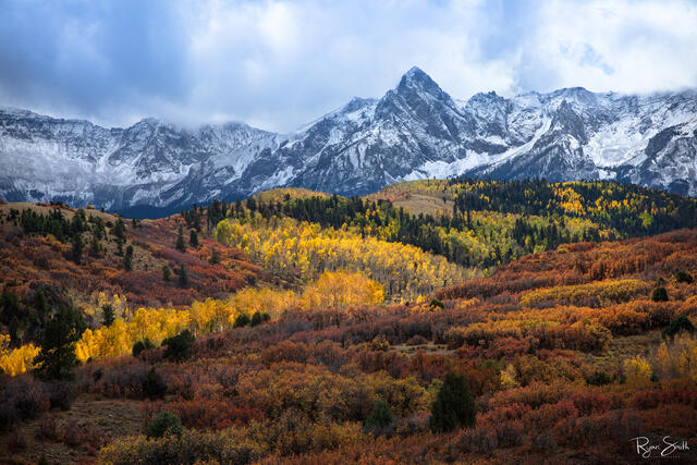 A snowy mountain skyline with white clouds in the sky stands with a valley full of Colorado fall color with yellow aspen trees, red and orange under brush,