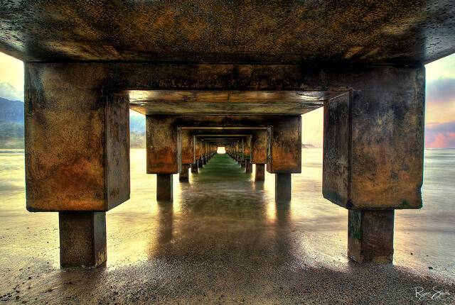 Symmetrical view from underneath a concrete pier seen at sunset with small square at the end of the pier glowing orange - a light at the end of the tunnel.