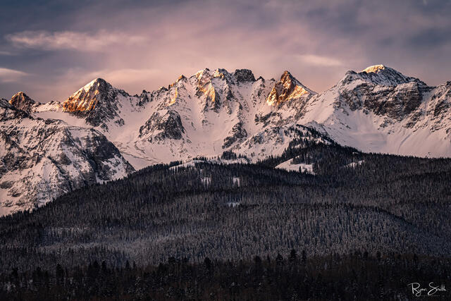 Tops of the snow covered mountains are hit with the first bit of sunlight with a snow dusted spruce trees surrounding the mountains.