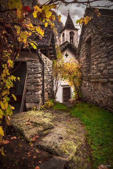 A small white Alpine chapel surrounded by weathered stone buildings and autumn foliage in a historic Swiss mountain village.