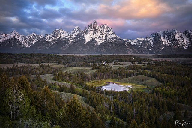 Grand Teton Mountain range stands tall in front of the sunset, under the pink clouds, and behind a landscape of water and green spruce trees.