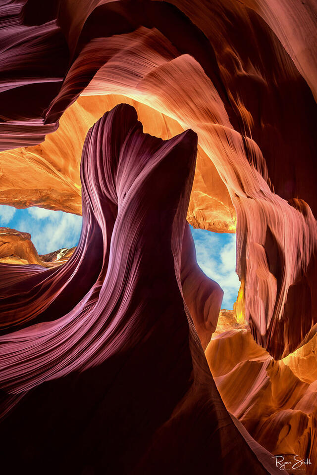 A slot canyon shows sandstone a pink rock formation with soft curves and lines with brighter orange canyon past it and a thin piece of blue sky with clouds.
