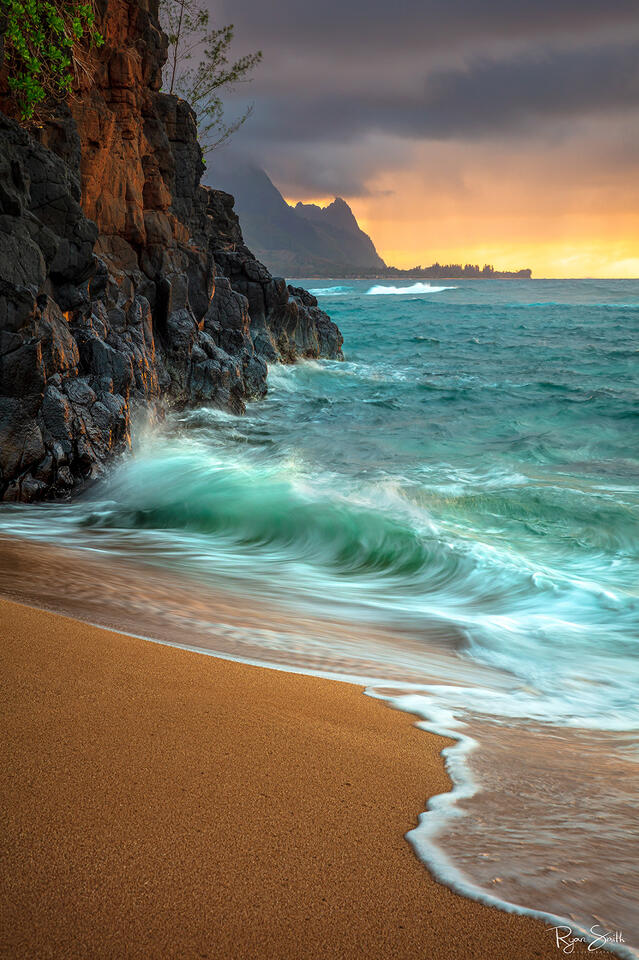 A shore is pictured in a vertical image with large rocks on the left and a vibrant orange sunset while aqua blue waves kiss the shore.