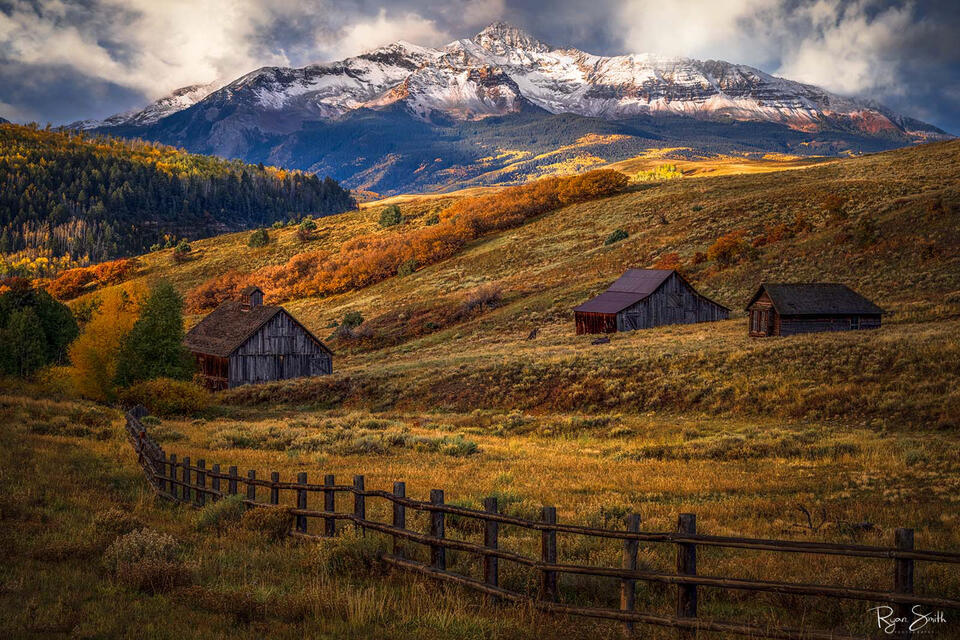 One cabin and two rustic barns sit on a hillside with the sun shining through the clouds, aspen trees in yellow, and oranges and mount wilson in the background.