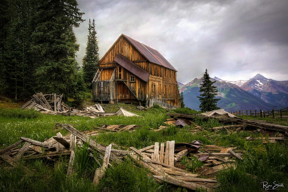 An abandoned wooden boarding house stands in a mountain meadow with dilapidated buildings and wooden boards throughout the green meadow.