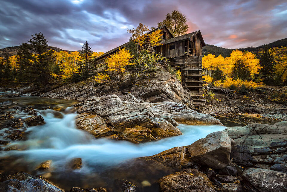 An old, rustic water mill sits in the Colorado hillside surrounded by bright yellow aspen trees and a few spruce trees with water flowing through the rocks.