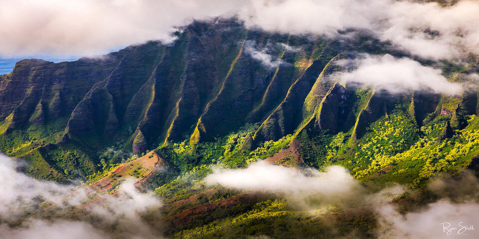 The sun lights up the valley of trees as clouds move through the lush green, tropical valley and along the top of the cliffs. 