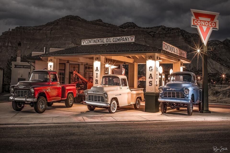 Three vintage Chevy trucks in red, white, & blue sit at a classic Conoco gas station with mountains and sky in the background in black and white as night falls.