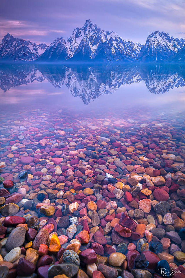 Colorful rocks sit below the cold, still water of the lake as it reflects the snow dusted mountains in the distance.