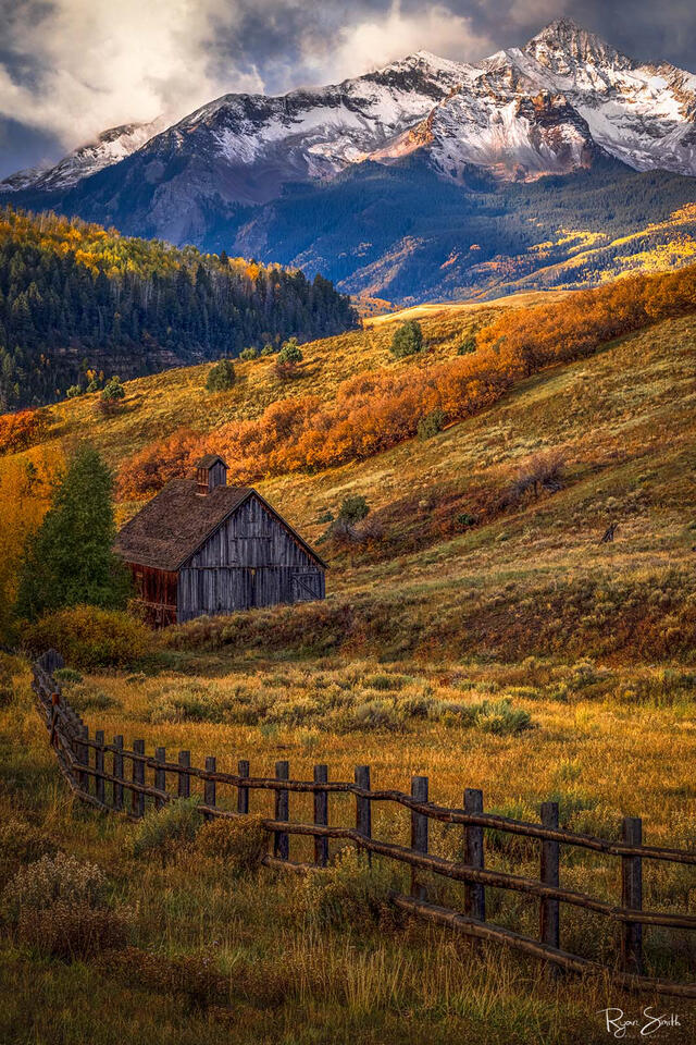 A rustic cabin sits on a hillside with the sun shining through the clouds, aspen trees in yellow, and oranges and mount wilson in the background with snow.