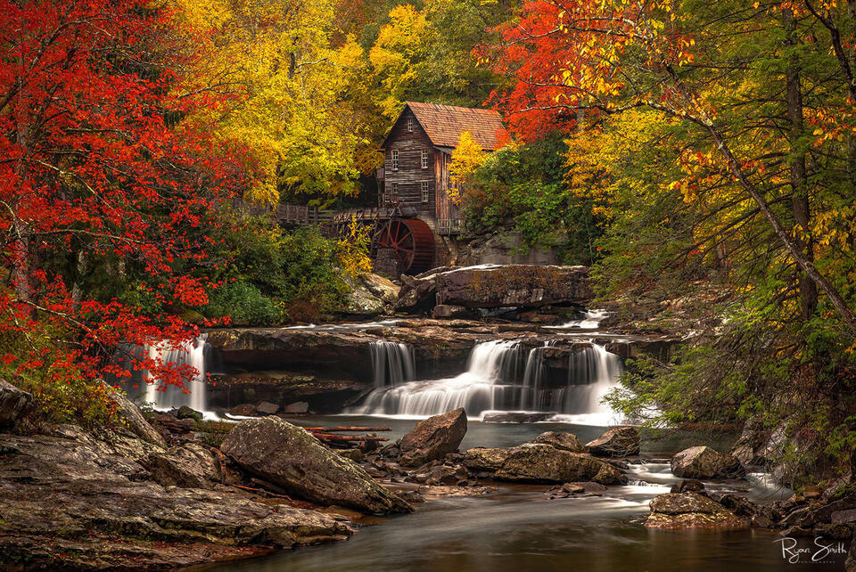 A rustic old mill sits on a mountain stream surrounded by fall color trees of yellow and flame red leaves.