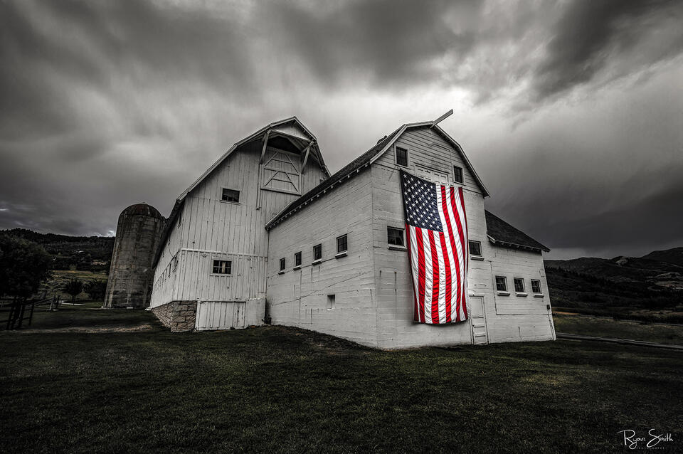 A dramatic black-and-white fine art photograph of the historic McPolin (Osguthorpe) Barn in Park City, Utah, with an American flag hanging proudly against its w