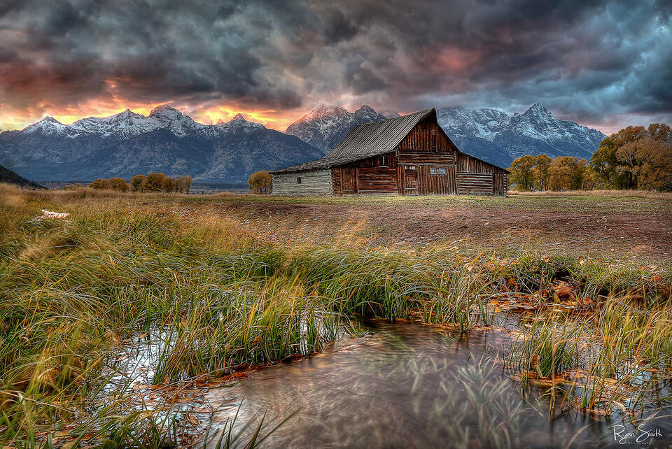 A rustic barn in front of a grand mountain view with clouds in the sky yet a break at the horizon shows a bright orange sky at sunset.