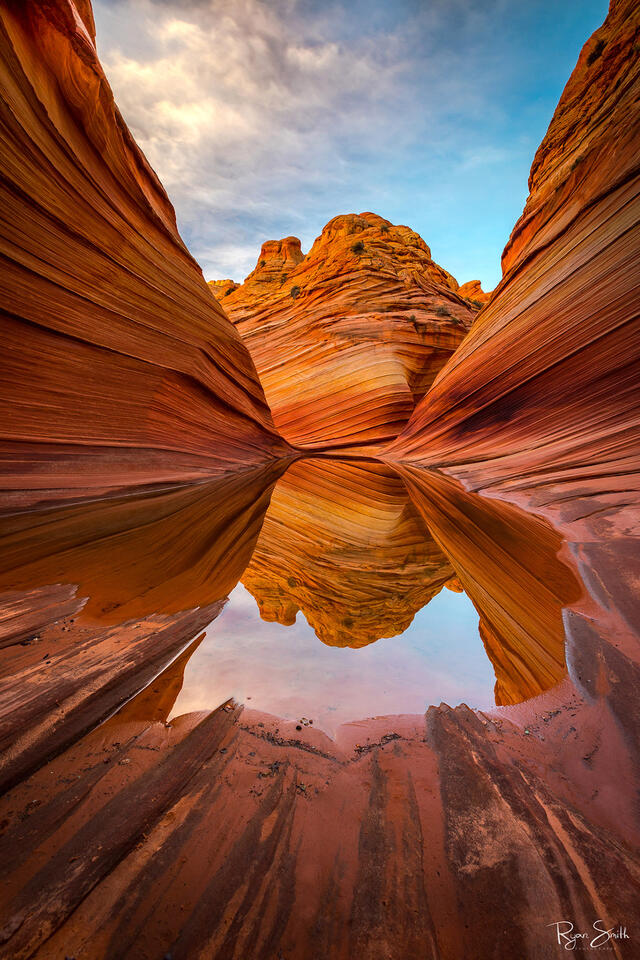 Sandstone walls with layers of red and orange colors are seen with a pool of water between them that reflects the rock formation at the far end of the photo.