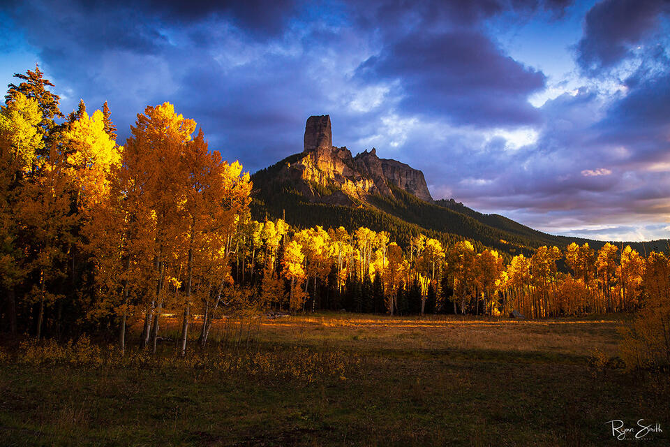 A straight, tall mountain resembles a chimney and towers above the spruce-covered hillside and meadow filled with bright yellow aspen trees. 