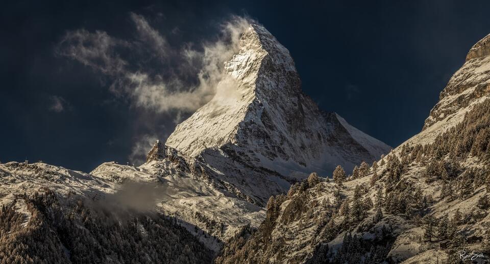 Fine art horizontal photograph of the Matterhorn in Zermatt, Switzerland with dramatic side light, wind-blown clouds, cool blue tones, and warm highlights.