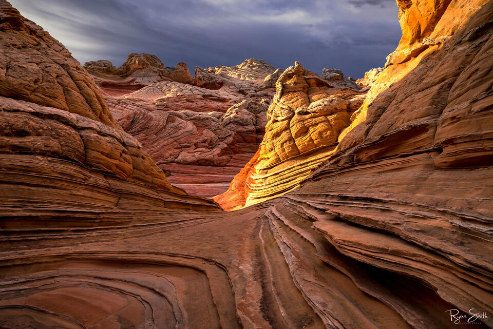 Layers of sandstone stack up the canyon walls while the sun sets and casts an orange glow on the edge of the wall with an overcast sky.