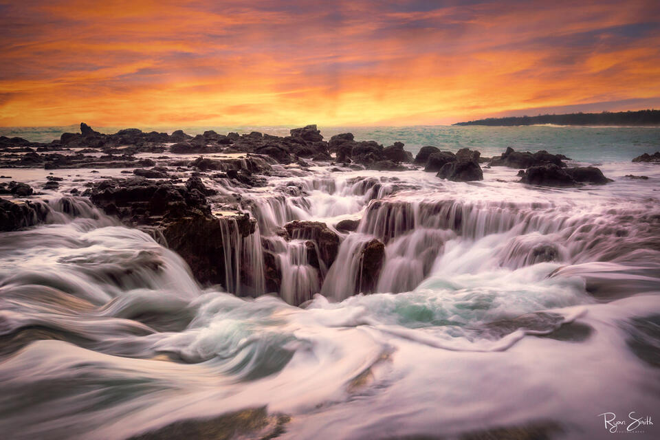 The sun sets on the distant horizon with a sky for yellow and orange while water rushes in and around a sinkhole on the rocky shore.