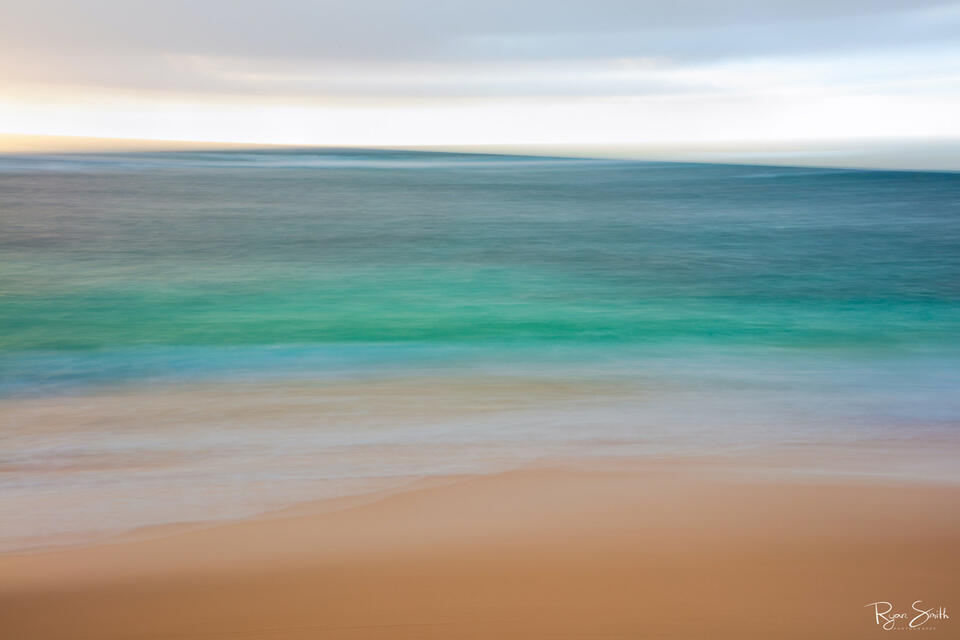 Abstract photography of the beach close up with the tan sand, shallow water and deep turquoise and blue water, and the cloudy, blurred sky.