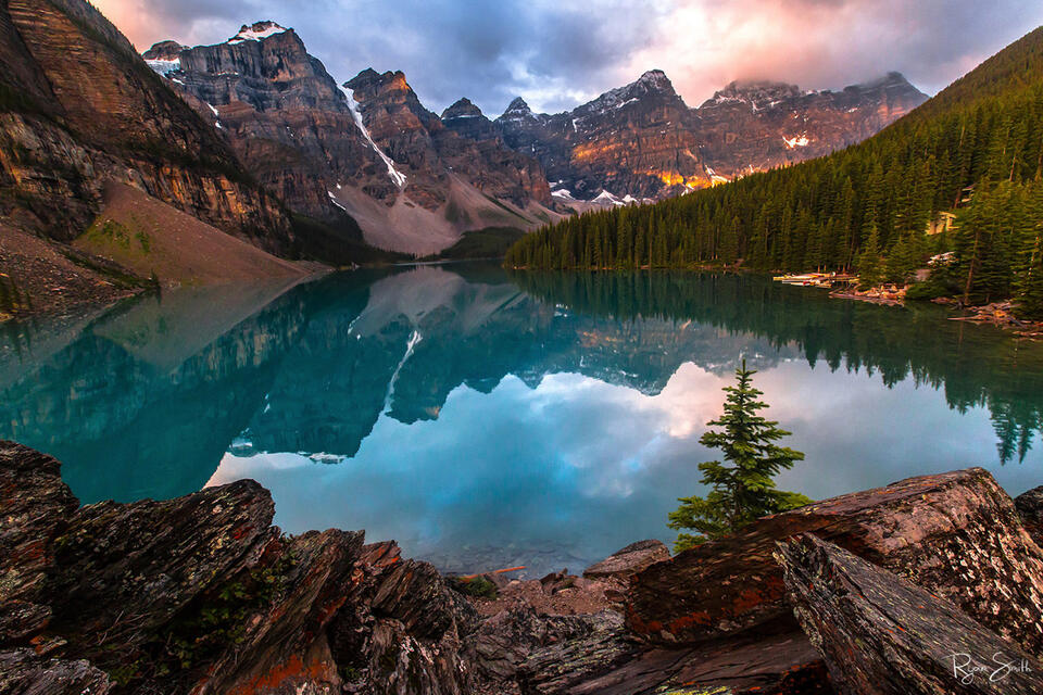 Turquoise blue lake reflects the sunlit mountain peaks, pink clouds, and spruce trees at sunrise in the Canadian Rocky Mountains