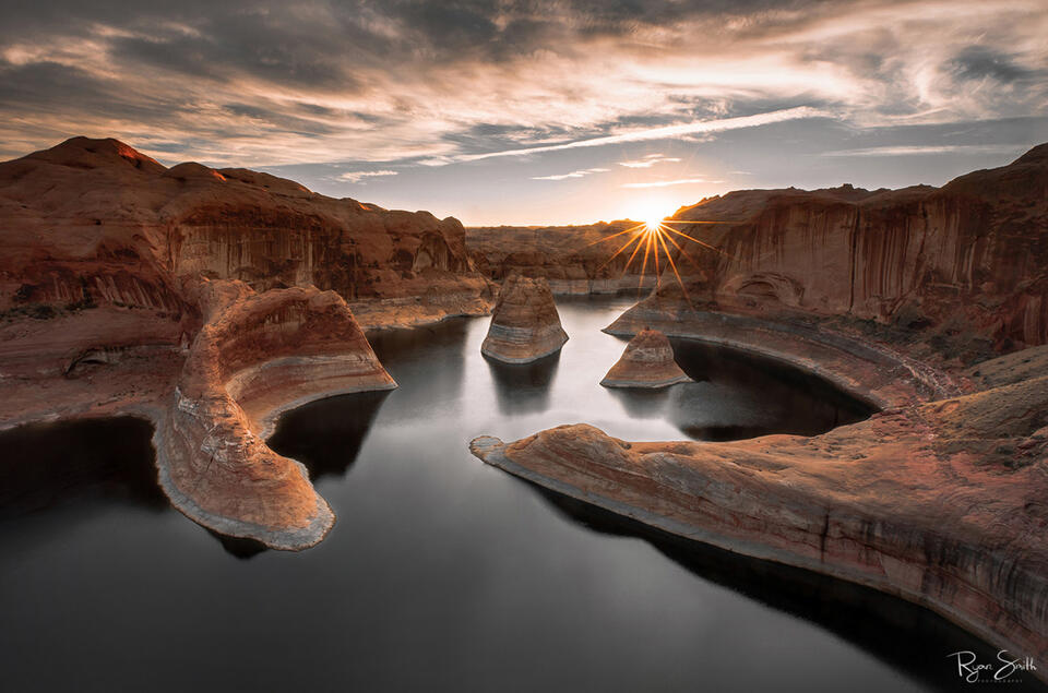Sunburst at sunrise shines new light on a still water winding through the pink and red walls of a canyon.