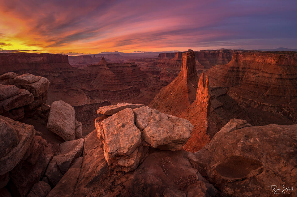 A canyon is lit at sunset with vibrant yellow, pink, and purple over the carved canviewpoint showing buttes and spires of pink and red layers rock.