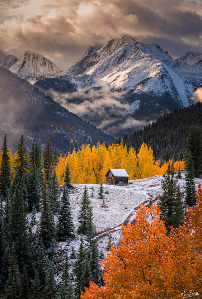 A picturesque scene featuring a grand mountain backdrop, vibrant fall colors, a dusting of snow, and a small quaint barn. A harmonious blend of seasons in one c