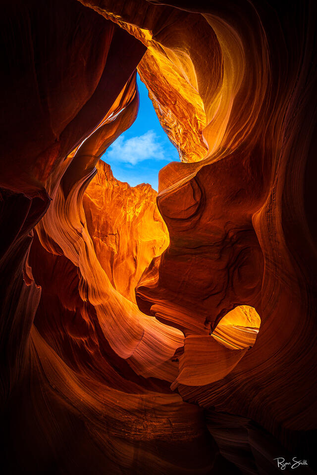 A slot canyon in shades of orange, red & yellow is shows as the sun shines light into the depths of the canyon & only a small triangle slot of sky is visible.