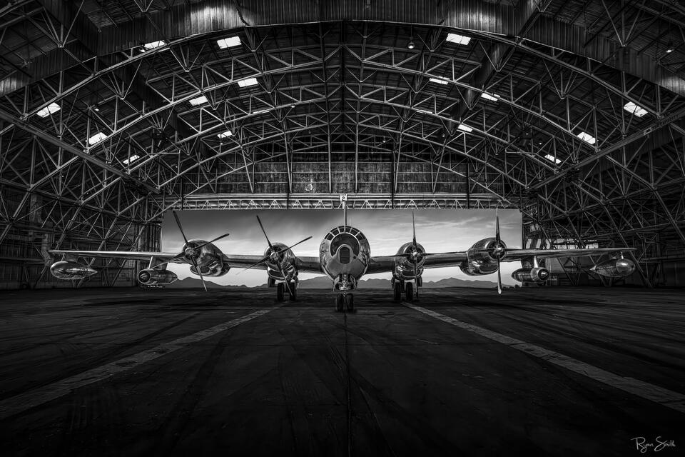 Black and white fine art photograph of a historic Superfortress-era aircraft resting inside a large steel aircraft hangar, emphasizing symmetry, industrial arch