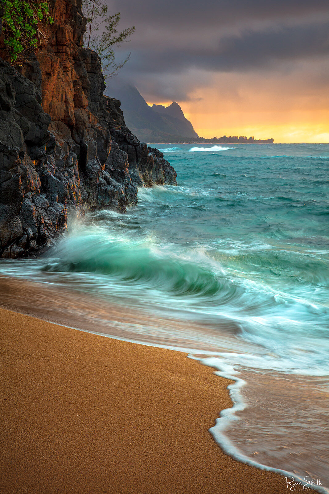 Hideaways A shore is pictured in a vertical image with large rocks on the left and a vibrant orange sunset while aqua blue waves kiss the shore.