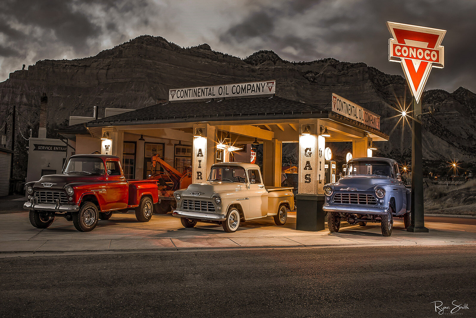 Three vintage Chevy trucks in red, white, & blue sit at a classic Conoco gas station with mountains and sky in the background in black and white as night falls.