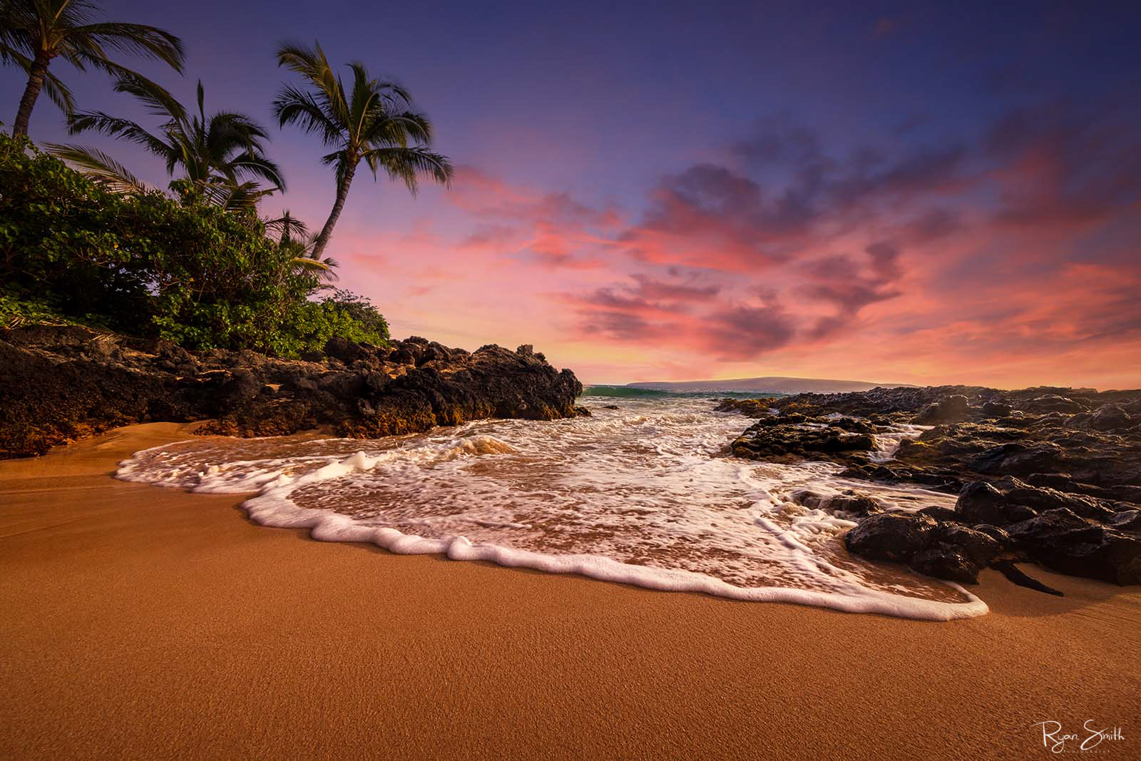 Sandy beach with volcanic rocks and palm trees at sunset as the sky is lit pink and the foamy white waves come in to kiss the shore. 