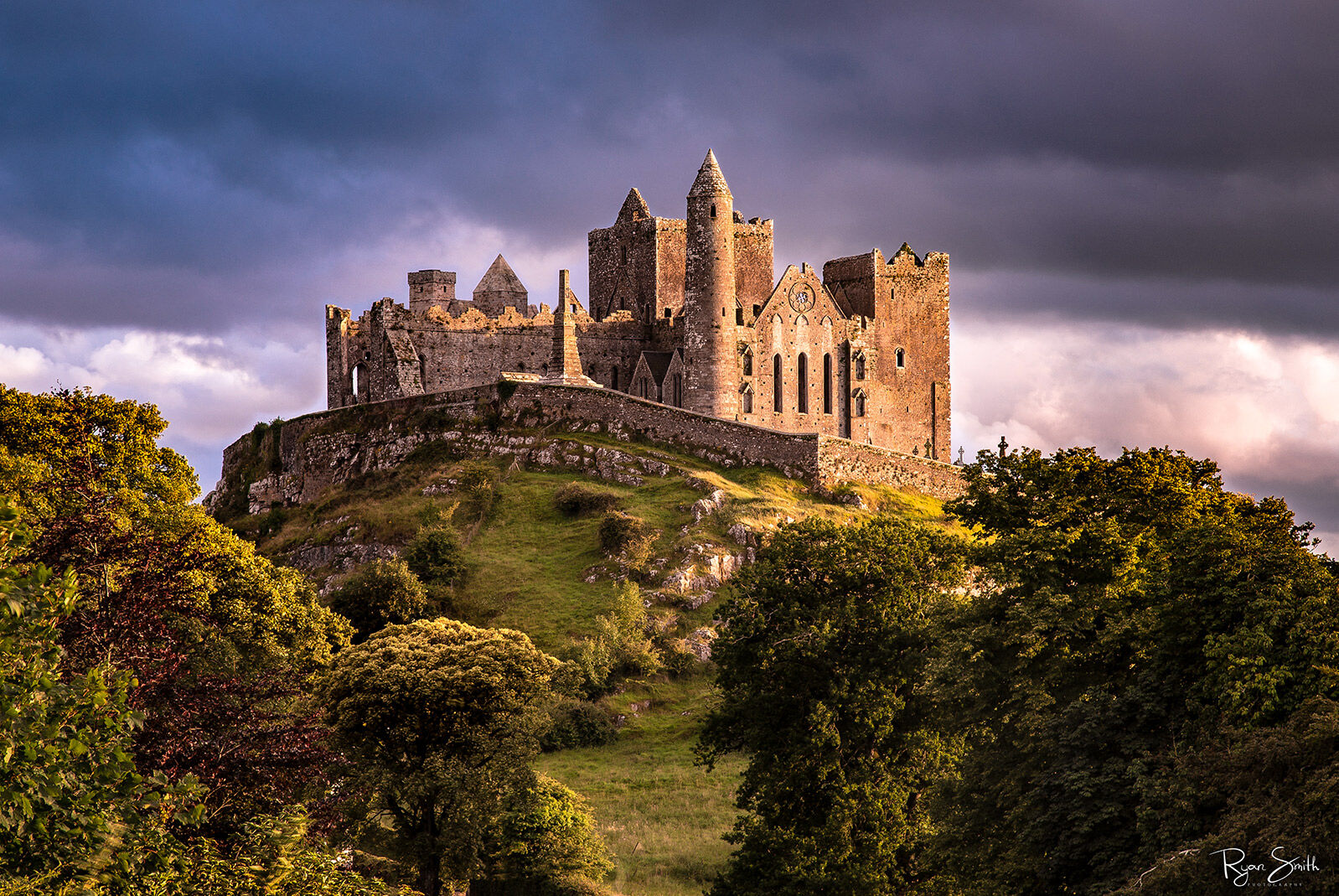 Rock of Cashel Castle A large castle sits up on a grassy hill with rocks and the sun setting behind it.