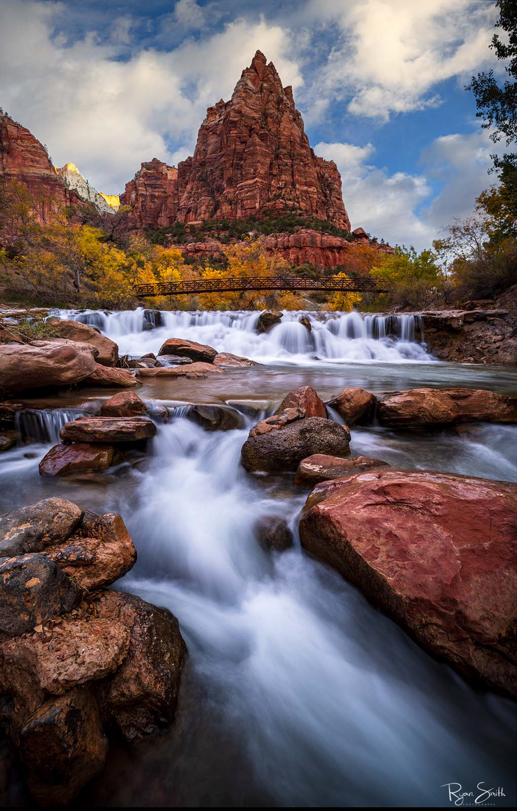 Zions Temple Jagged mountain with a well defined peak stands in front of a blue sky with white clouds with a bride and waterfalls pouring down the front of the scene.