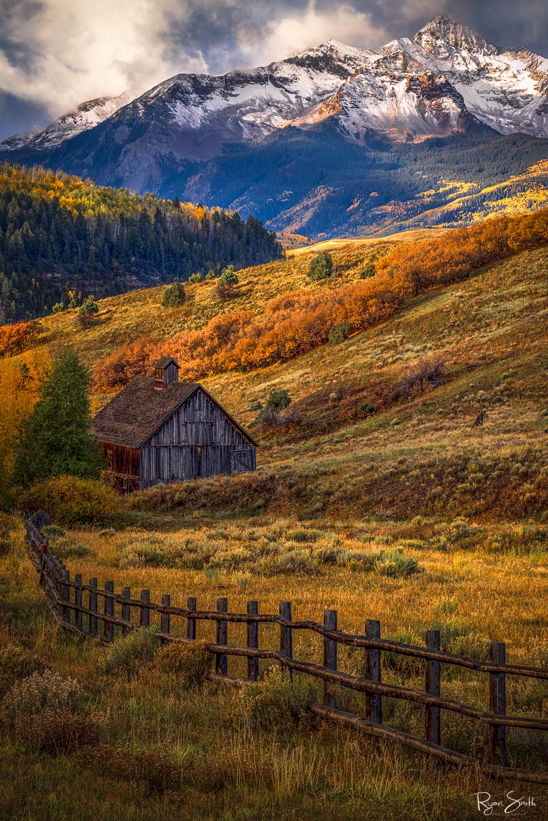 Colorado Homestead A rustic cabin sits on a hillside with the sun shining through the clouds, aspen trees in yellow, and oranges and mount wilson in the background with snow.