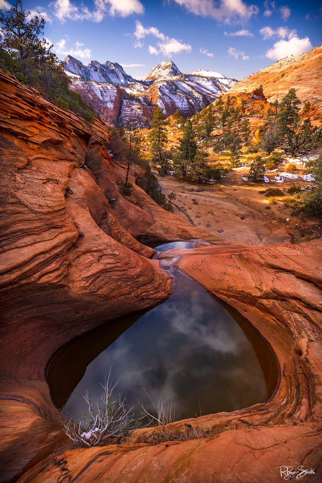 Snowy mountains under clouds in a blue sky while the sun lights up the hillside as new life grows and a reflection pool surrounded by red rock mirrors the sky.
