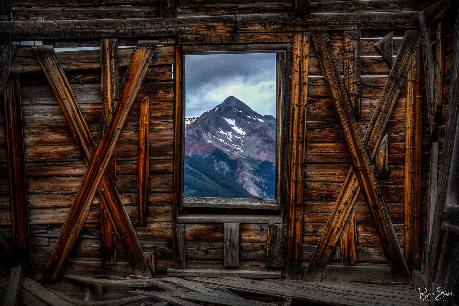 Inside an abandoned cabin, the floor boards have fallen but the wall still stands and the paneless window reveals a grand mountain view.