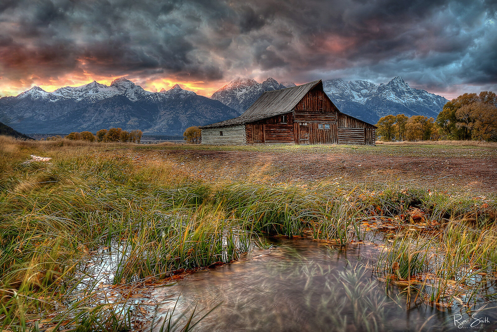 Teton Nightfire A rustic barn in front of a grand mountain view with clouds in the sky yet a break at the horizon shows a bright orange sky at sunset.