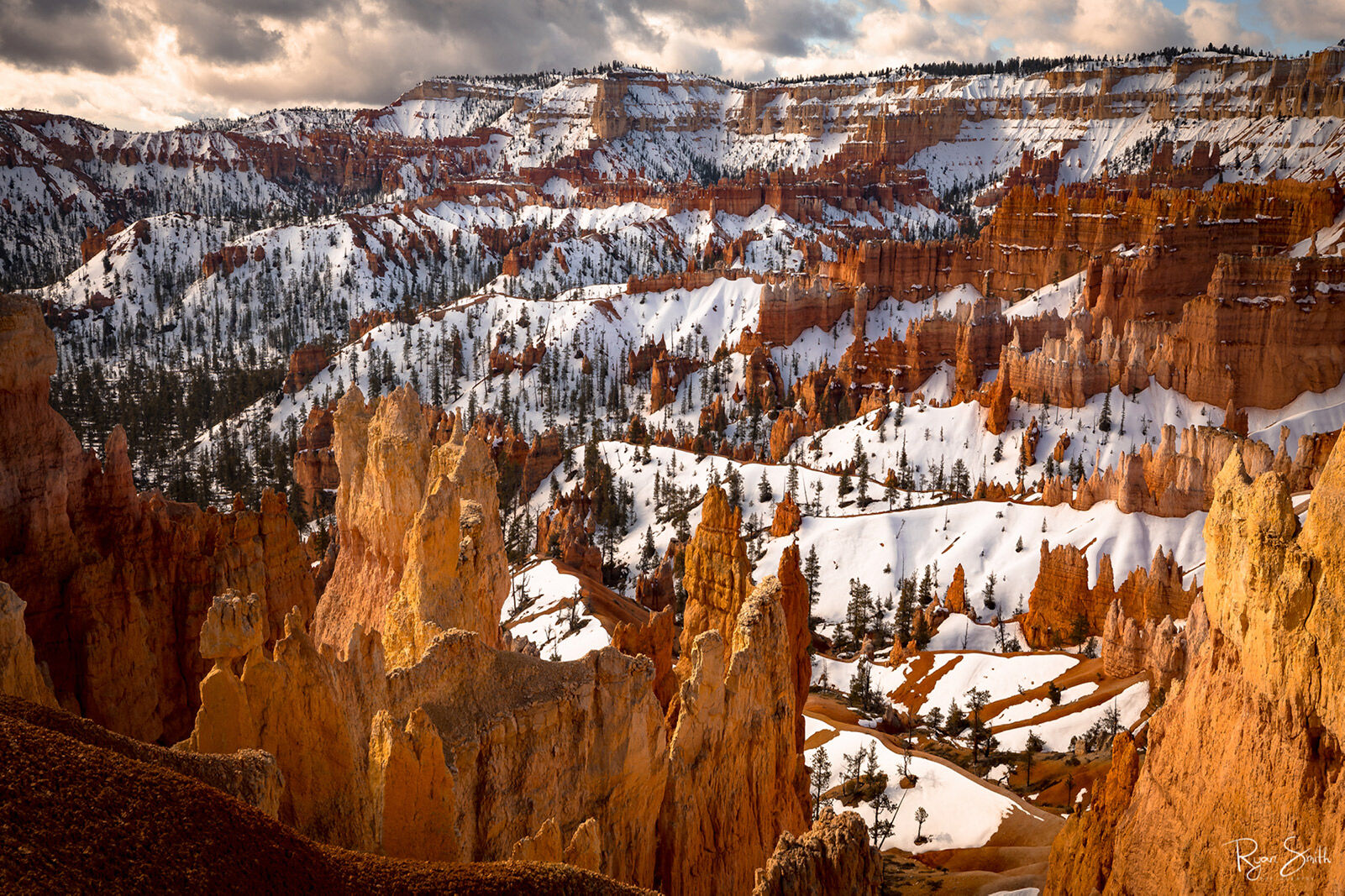 A canyon filled with red hoodoos, or rock spires, is seen with snow over the landscape and some trees in the canyon and the sky filled with fluffy clouds.