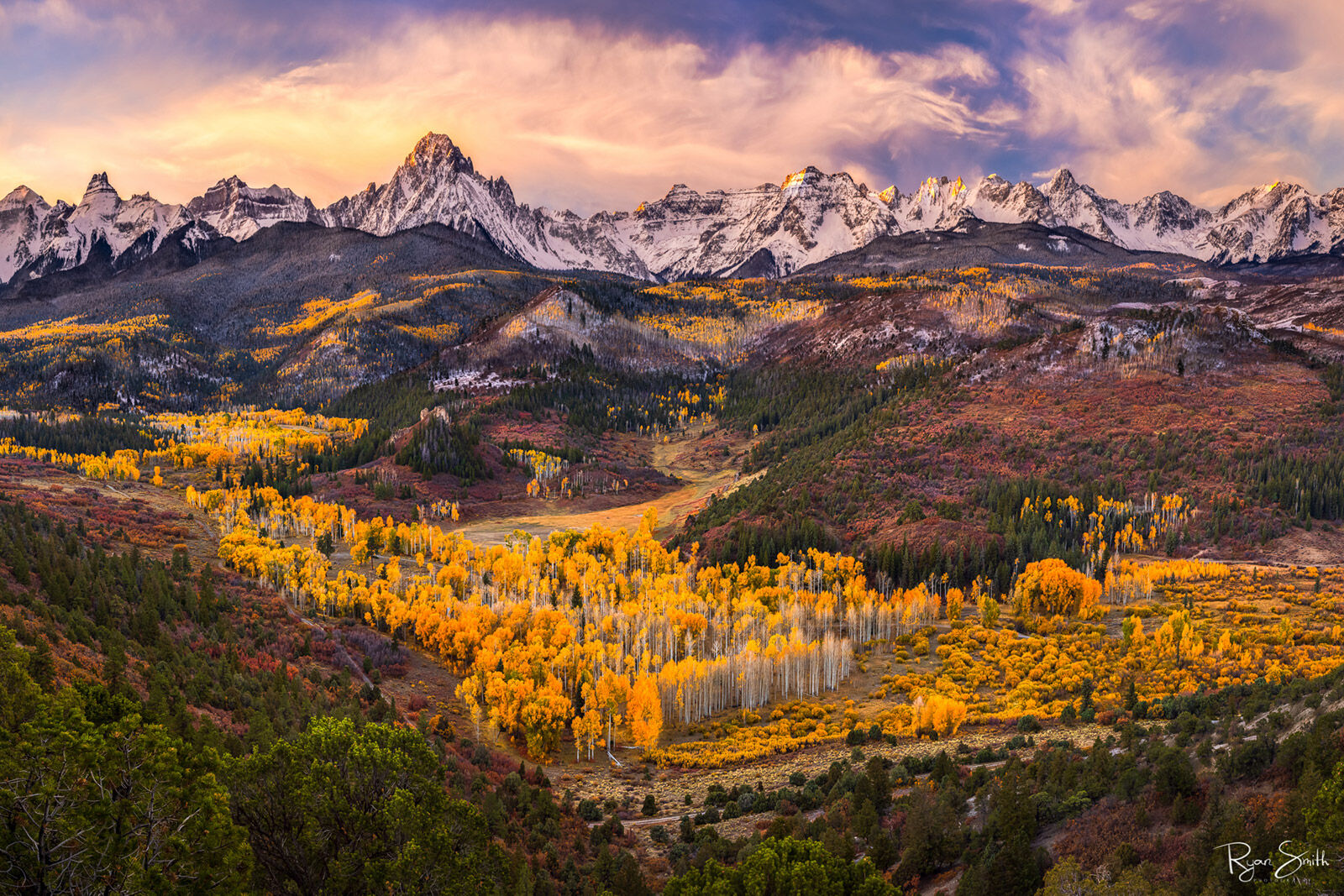 Mountain skyline covered with snow with a valley of gold leaved aspen trees as the setting sun light just touches the tops of the mountains & lights the clouds
