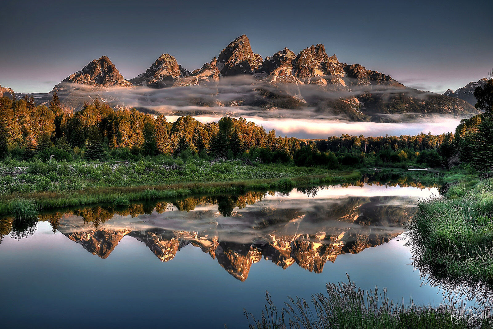 Mountain skyline is seen at sunrise with thin clouds in below the peaks. A lake reflects the mountains, clouds & spruce trees that sit in front of the mountain.