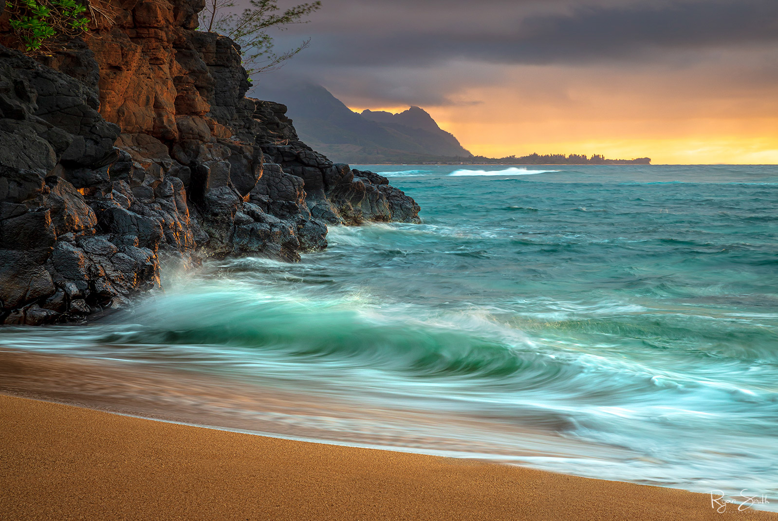 A shore is pictured with large rocks on the left and a vibrant orange sunset while aqua blue waves kiss the shore. 