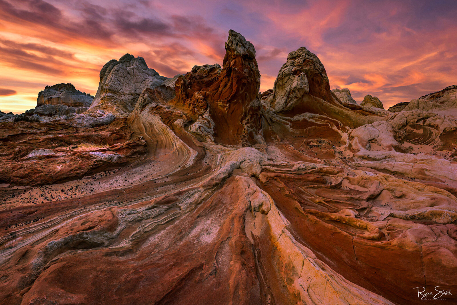 A landscape of tall rocks show signs of being worn away over the years with grooves worn by water. Shades of pink and deep reds in the rocks and pink in the sky