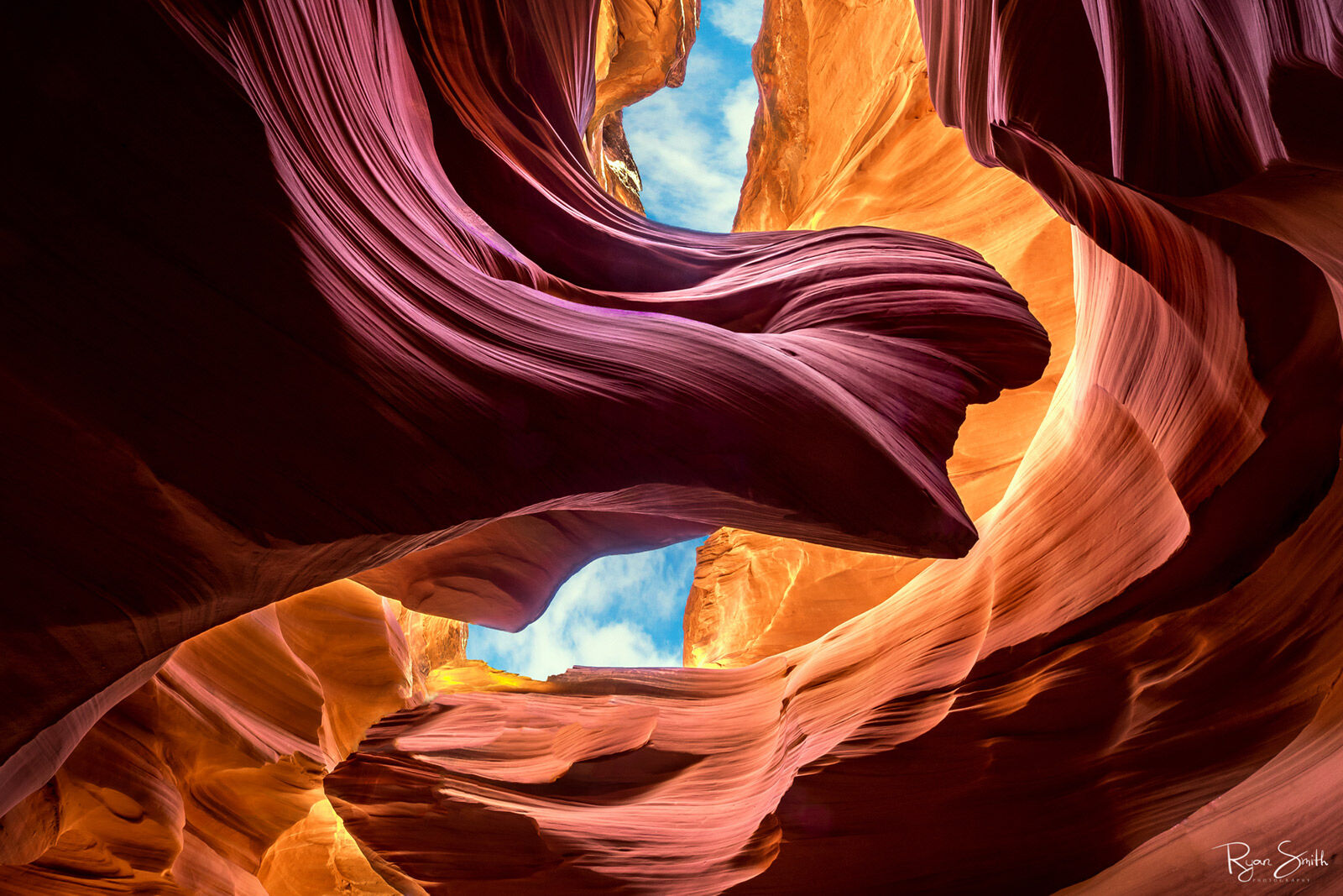 Inside a slot canyon are rock formations & walls that have fine lines and curves carved by wind and water with a small piece of blue sky & white clouds visible.