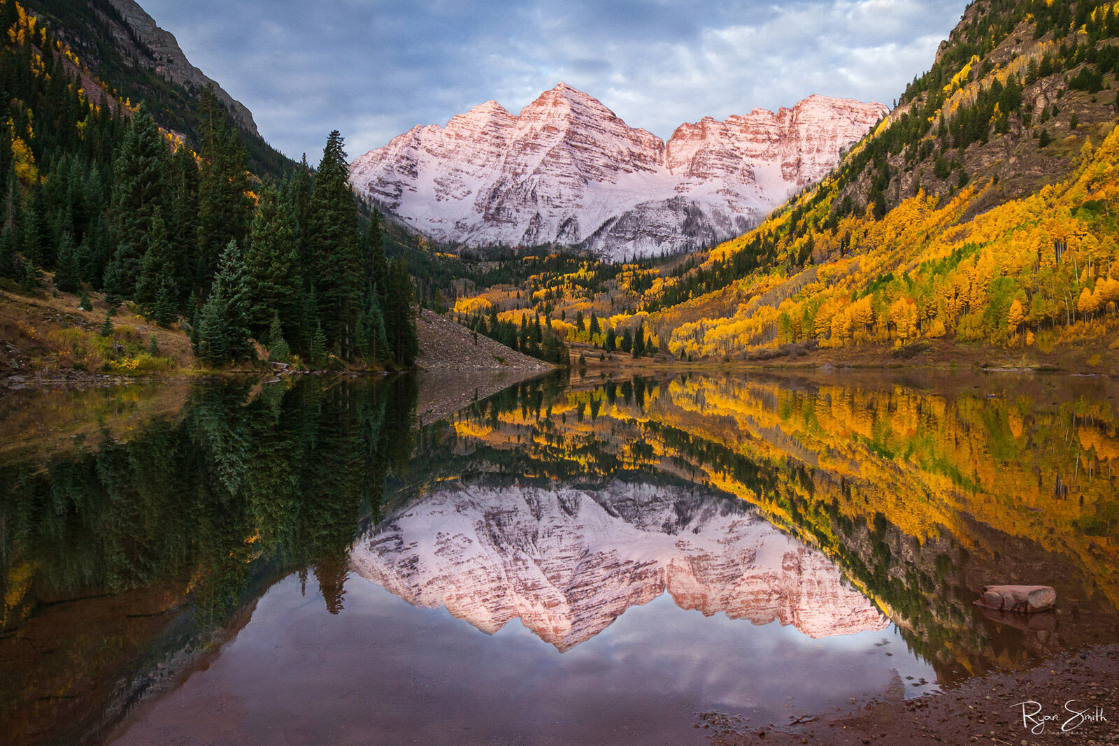 Snow covered mountains are touched by the sunlight and surrounded by hillsides of fall colors with the whole scene reflected in the still lake. 