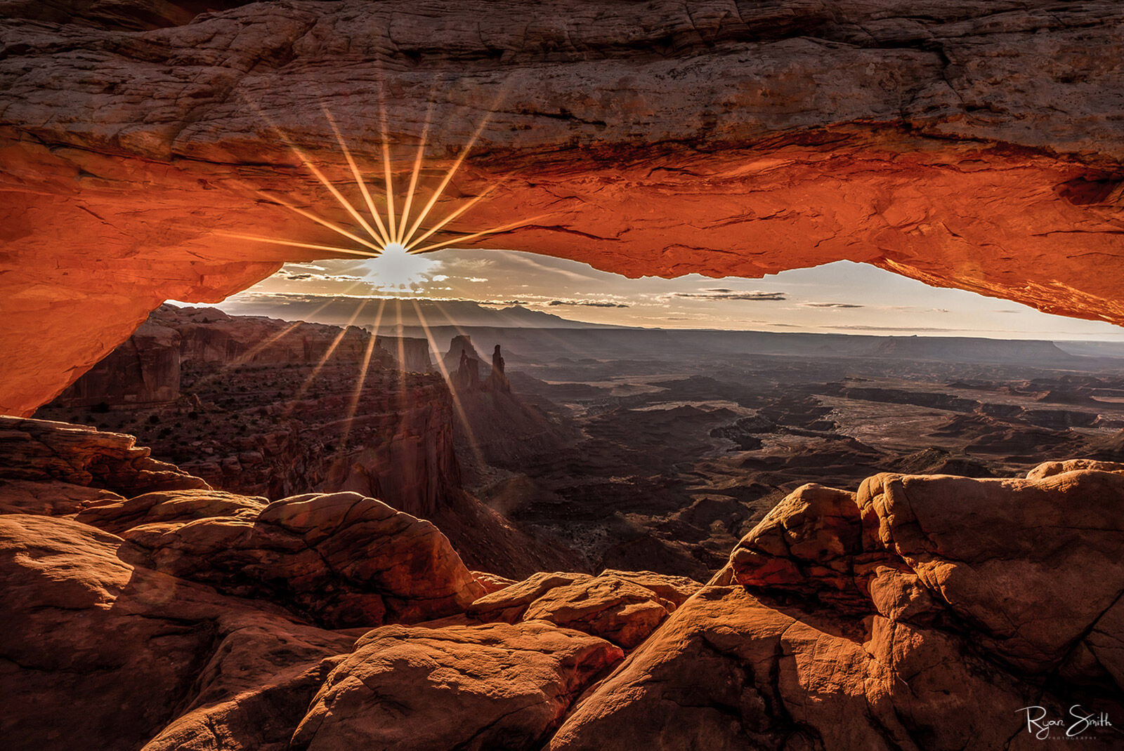 The sun rises and is viewed from under a sandstone arch that glows orange as the canyon below can be viewed and a sunburst sparkles from under the arch.