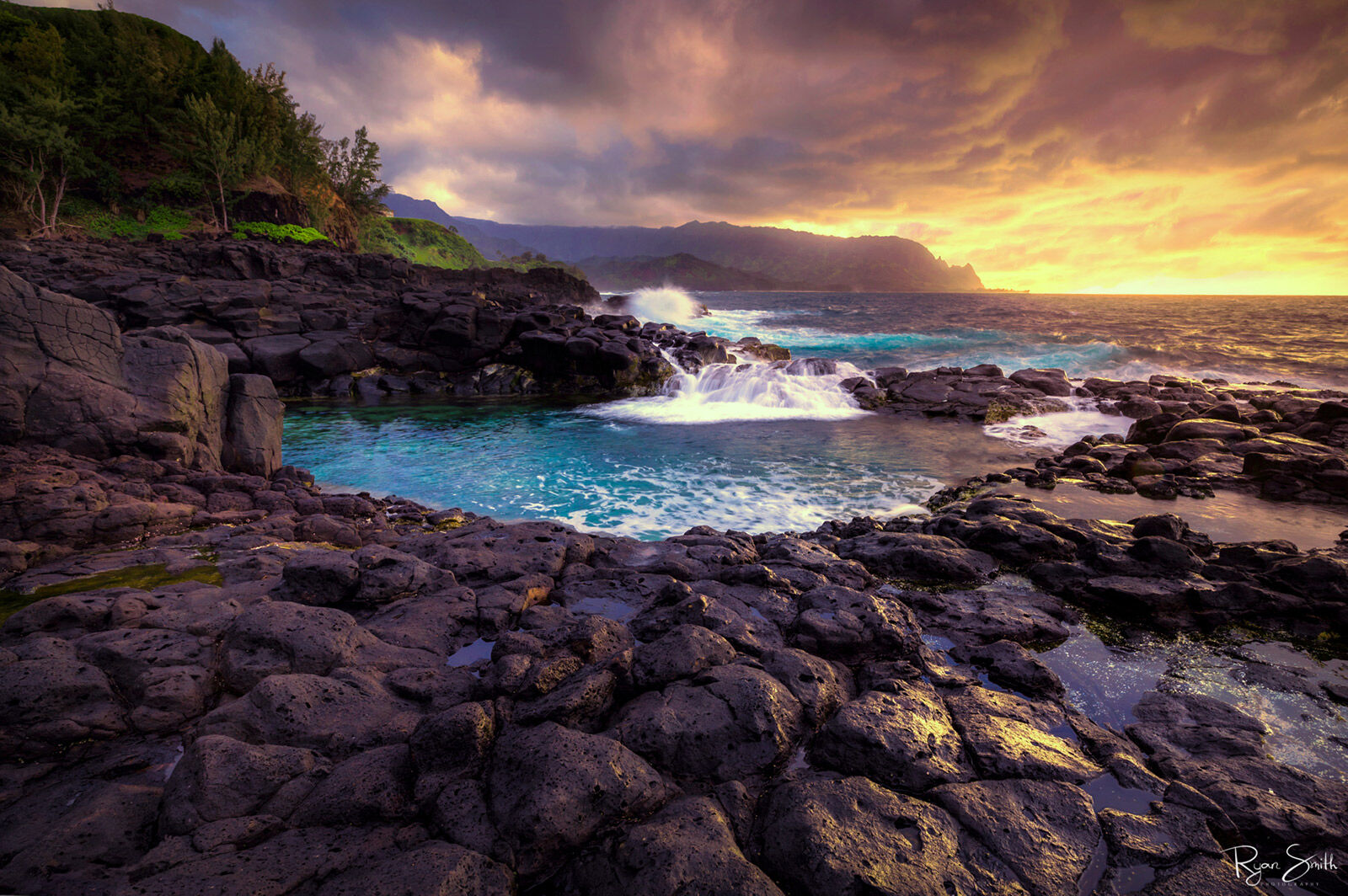 A rocky shore has a round pool with the ocean flowing in and the sun setting in the distance with an orange cloudy sky.