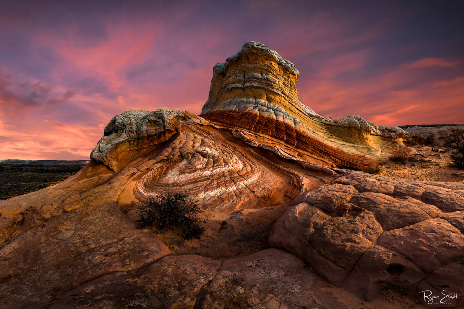 Deep orange, yellow, pink, and white layes of a rock formation rise above the desert landscape while shades of pink and blue hang in the sky at sunset.